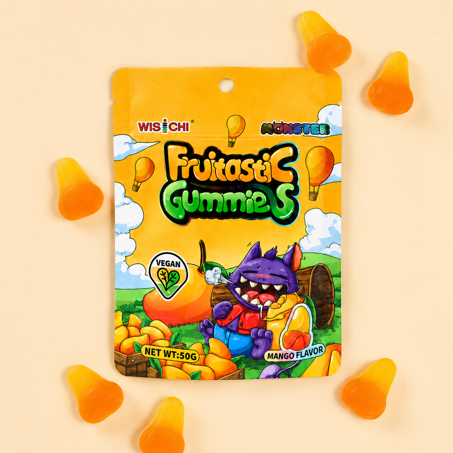 A 50g bag of Wisichi Minster Fruitastic Gummies (Mango Flavor) displayed on a light beige background with scattered orange and yellow gummies around it. The packaging features a bright yellow-orange background with cartoon graphics of a purple monster, a mango, and hot air balloons.
